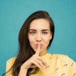 portrait photo of woman in yellow t shirt doing the shh sign while standing in front of blue background